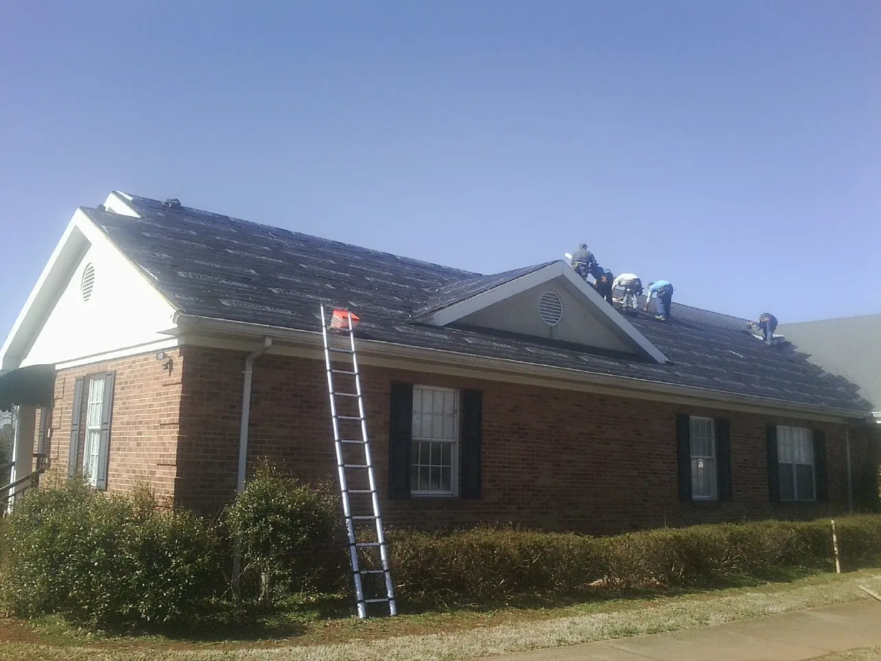 Crew tearing shingles off a brick ranch roof with a ladder set — Emergency Roof Repair work by our crew in Dallas Center
