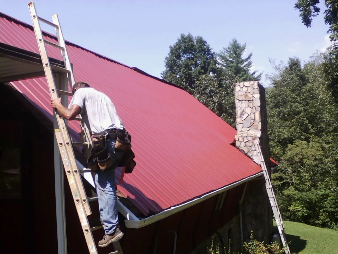 Roofer on a ladder beside a red metal roof and stone chimney — Soffit & Fascia Repair by Roofing Brothers in Dallas Center